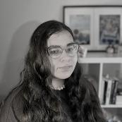A young woman with dark hair and eyes with artwork on the wall behind her.